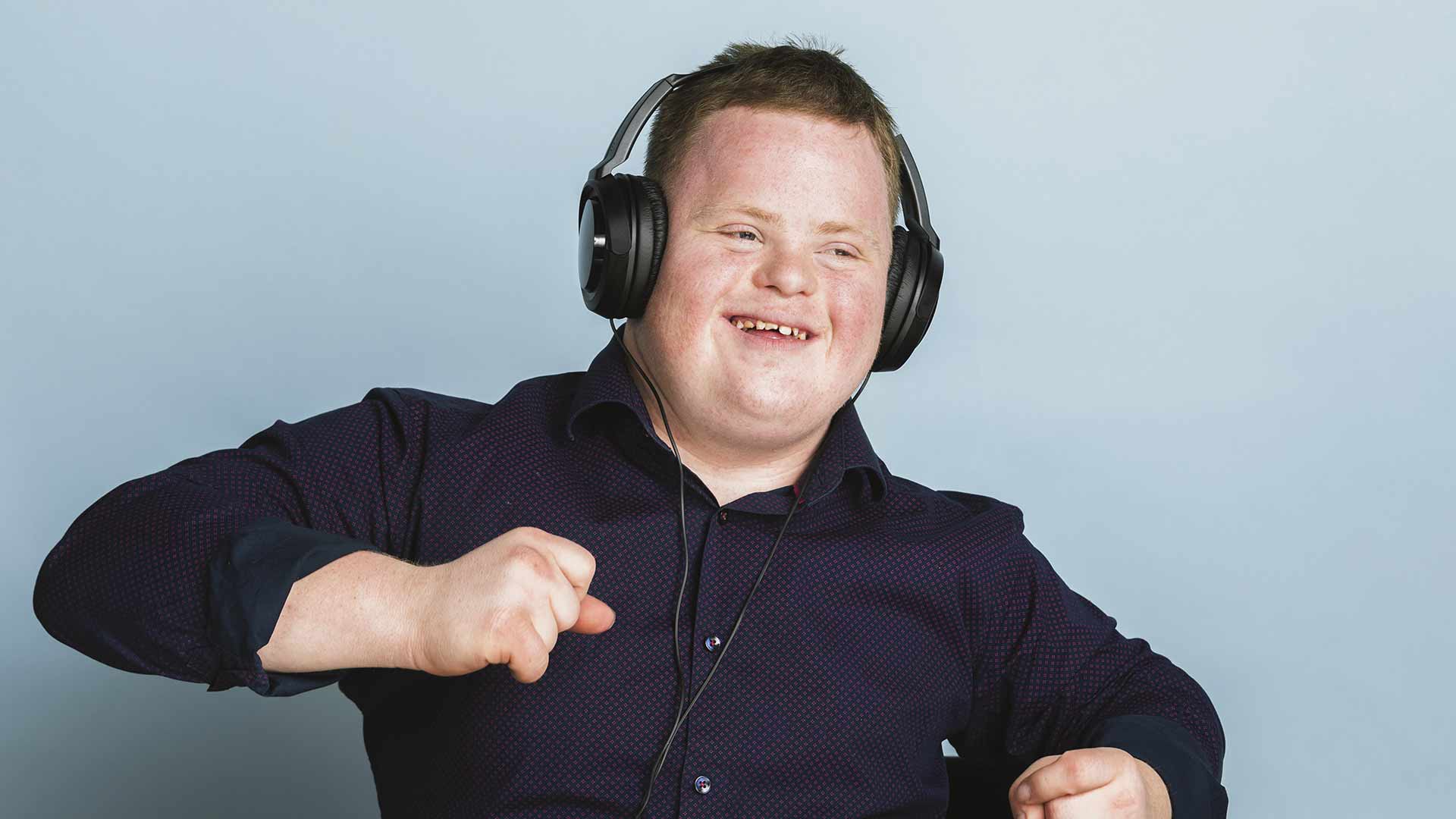 A young man with downs syndrome dances in front of a blue wall with headphones on